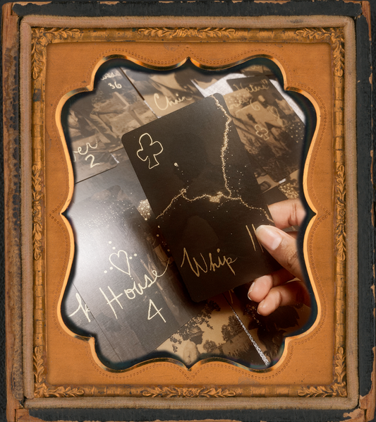 A brown hand holds the black and gold Whip card from Grandma Baby's Black Gold Lenormand Too. in the background the other cards lay face up in neat rows and columns. this photo sits inside a vintage brass picture frame.