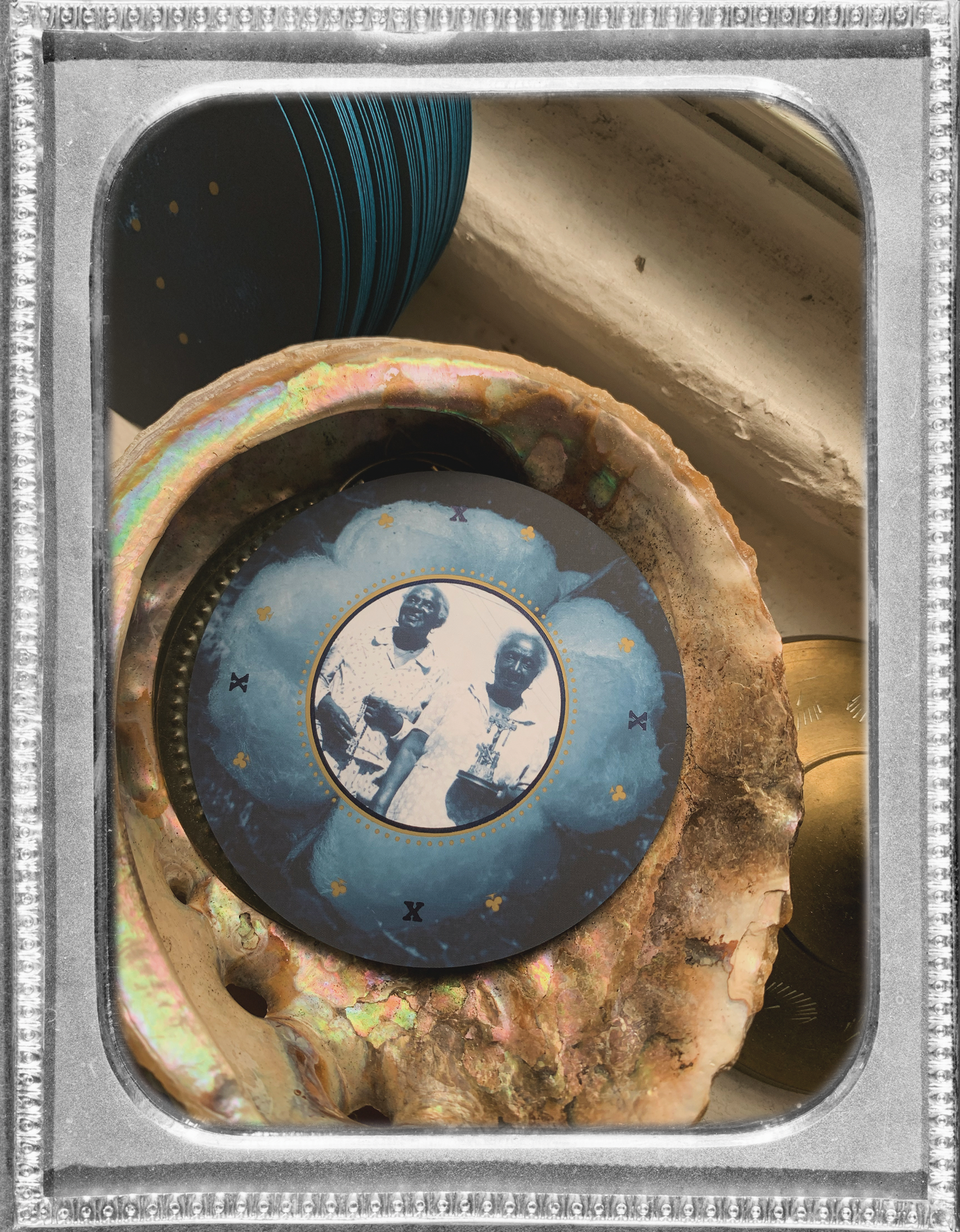 inside a silver vintage frame is a photo of a circular playing card from Grandma Baby’s 52 Blues Playing Cards deck laying face up inside an abalone shell. the other cards in the deck are laying beside the shell on a white windowsill, tilted to reveal their blue edges.