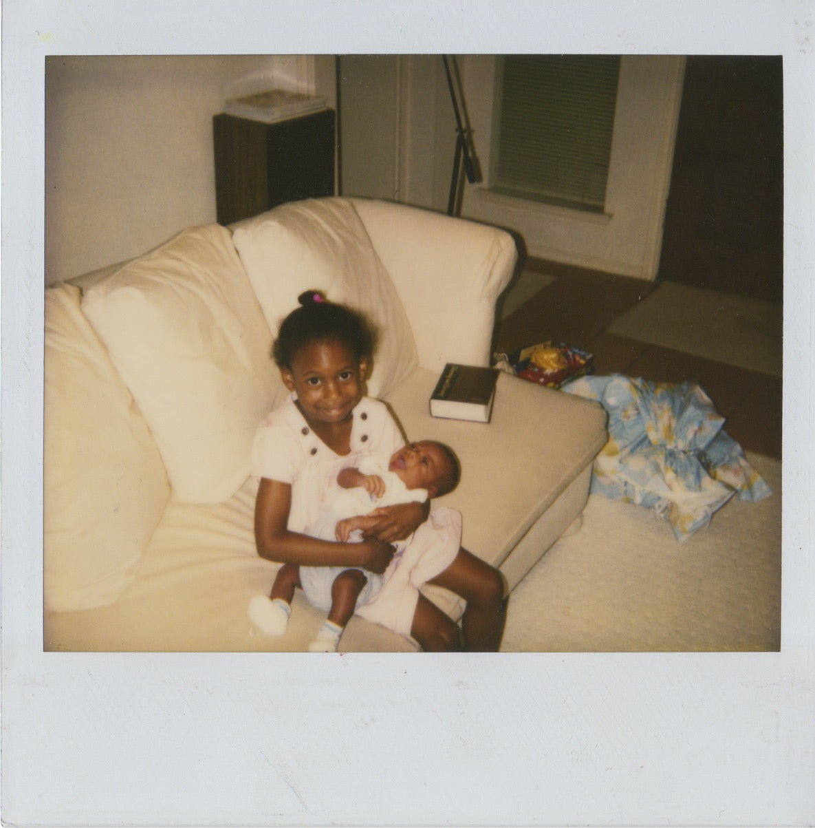 old polaroid photograph of two young Black children, one a toddler and one an infant cradled in the toddlers arms.