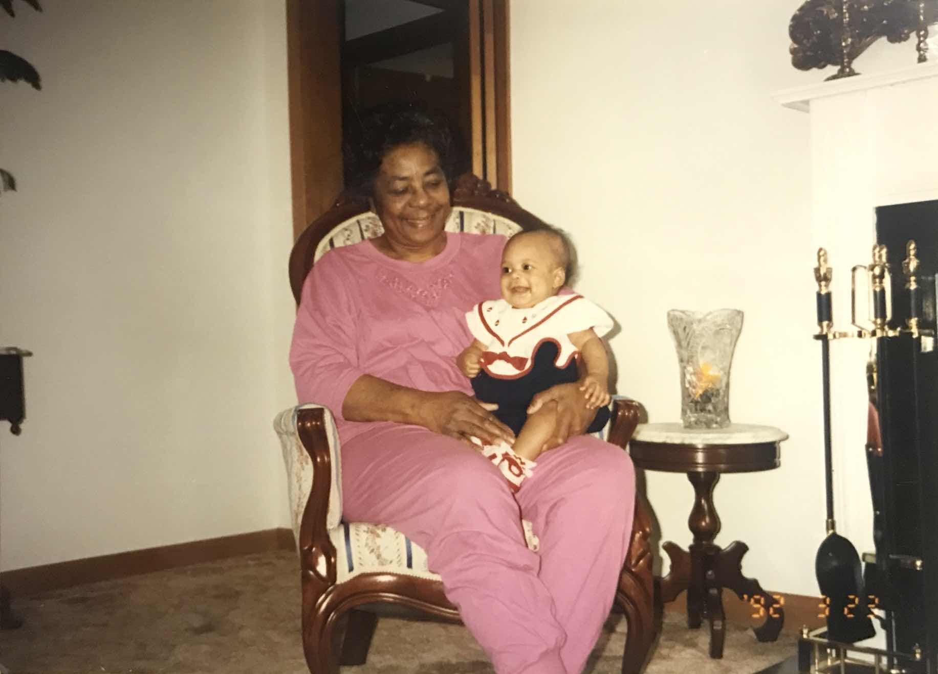 old photograph of a Black grandmother in a pink lounge set holding her infant grandchild. They are both smiling.