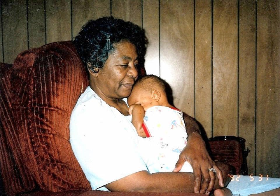 Old photograph of a Black grandmother in a light blue house dress seated in a red recliner, holding her infant grandchild to her chest.