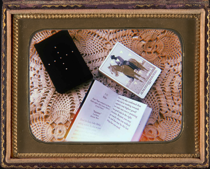 On top of a white crocheted blanket are the black and gold tuckbox, the open guidebook and the cards from the Grandma Baby's Black Gold Lenormand deck. the guidebook is open to the entry for the Rider card and the card laying face up on top of the deck is also the Rider card. this photo is inside a vintage gold or brass picture frame.