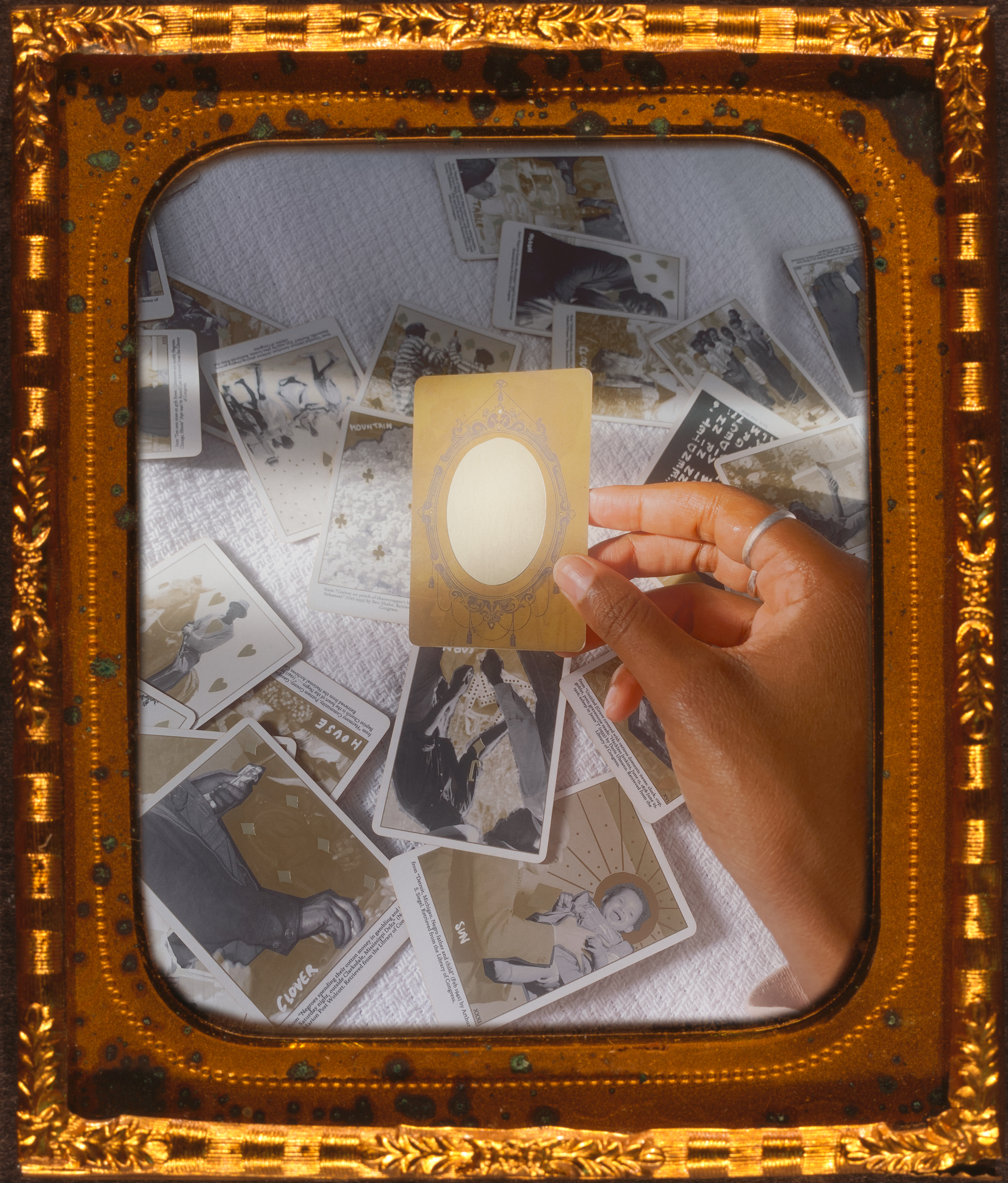 a brown hand holding the Mirror card from the Grandma Baby's Black Gold Lenormand deck above a white bedspread covered in the remaining cards from the deck. a sliver of natural sunlight falls across the cards, reflecting off the surface of the Mirror card. this photo is inside a brass or gold vintage picture frame.