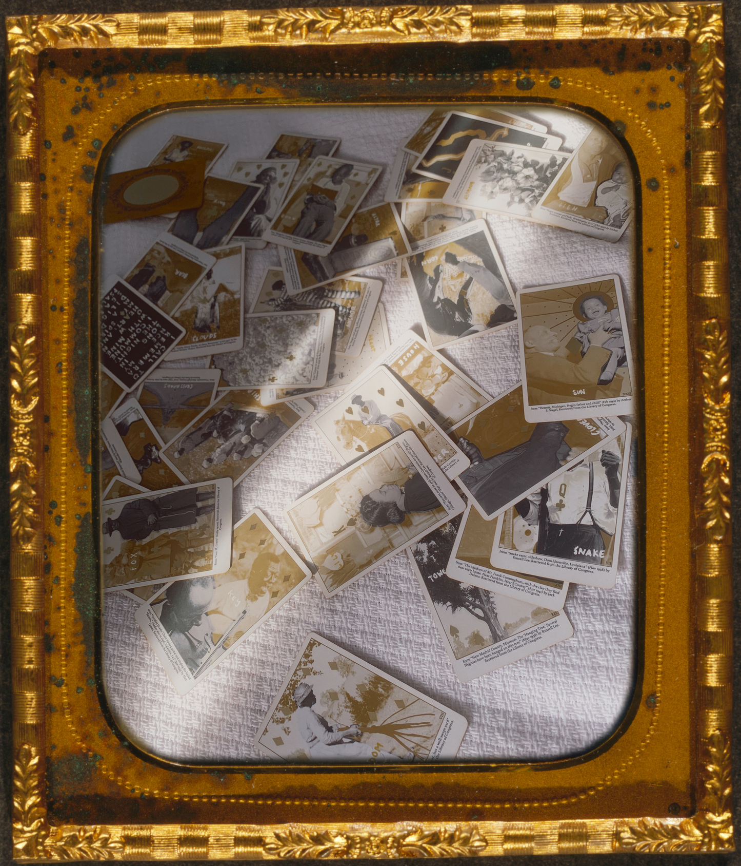 a bronze daguerreotype frame around a photo of the cards from Grandma Baby’s Black Gold Lenormand deck scattered face up across a white bedspread as a sliver of sunlight slashes across them.