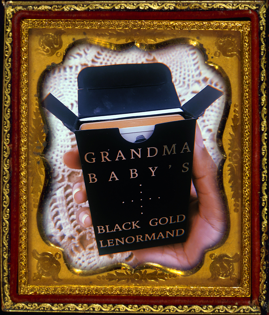 a brown hand holds the Grandma Baby's Black Gold Lenormand deck with the black and gold tuck box open, revealing the slim guidebook and matte gold edges of the cards. in the background there is a white crocheted blanket. this photo is inside a vintage brass picture frame.