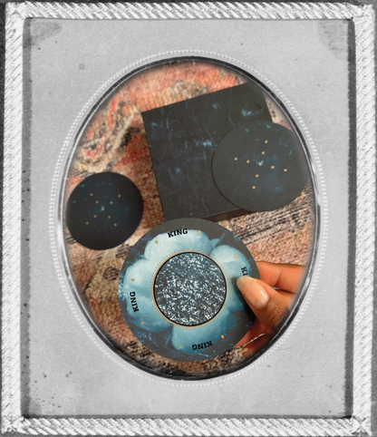 inside a vintage silver frame, a photo of a brown hand holding a circular blue playing card in the foreground while more cards and the deck box are laying in the background